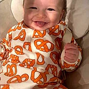 Luca is registered to the contest to win money with this photo: baby, blanket, child, clothing, cozy, cushion, face, happy, indoor, infant, onesie, orange, patterned_clothing, photography, pillow, portrait, pretzel, seated, smiling, white