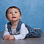 baby, child, infant, portrait, overalls, white_shirt, floor, blue_background, curious, cute, lying_down, hands_clasped, studio_photo, young_child, face, eyes, hair, indoor, clothing, expression