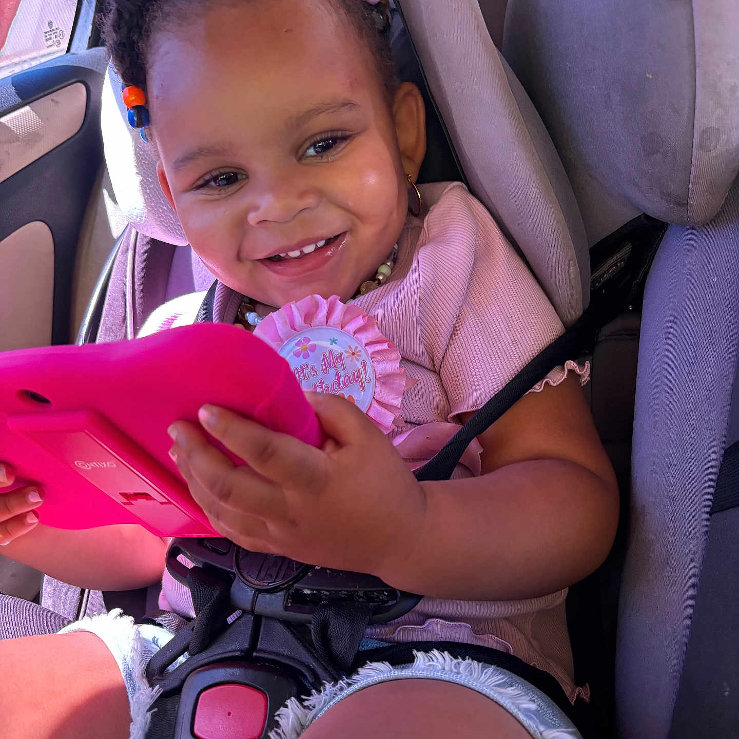 Nevaeh is registered to the contest to win money with this photo: toddler, child, car_seat, birthday_badge, tablet, smiling, hair_beads, summer_clothes, casual_wear, sunlight, car_interior, seatbelt, happy, cute, portrait, close_up, baby_girl, playful, technology, fun
