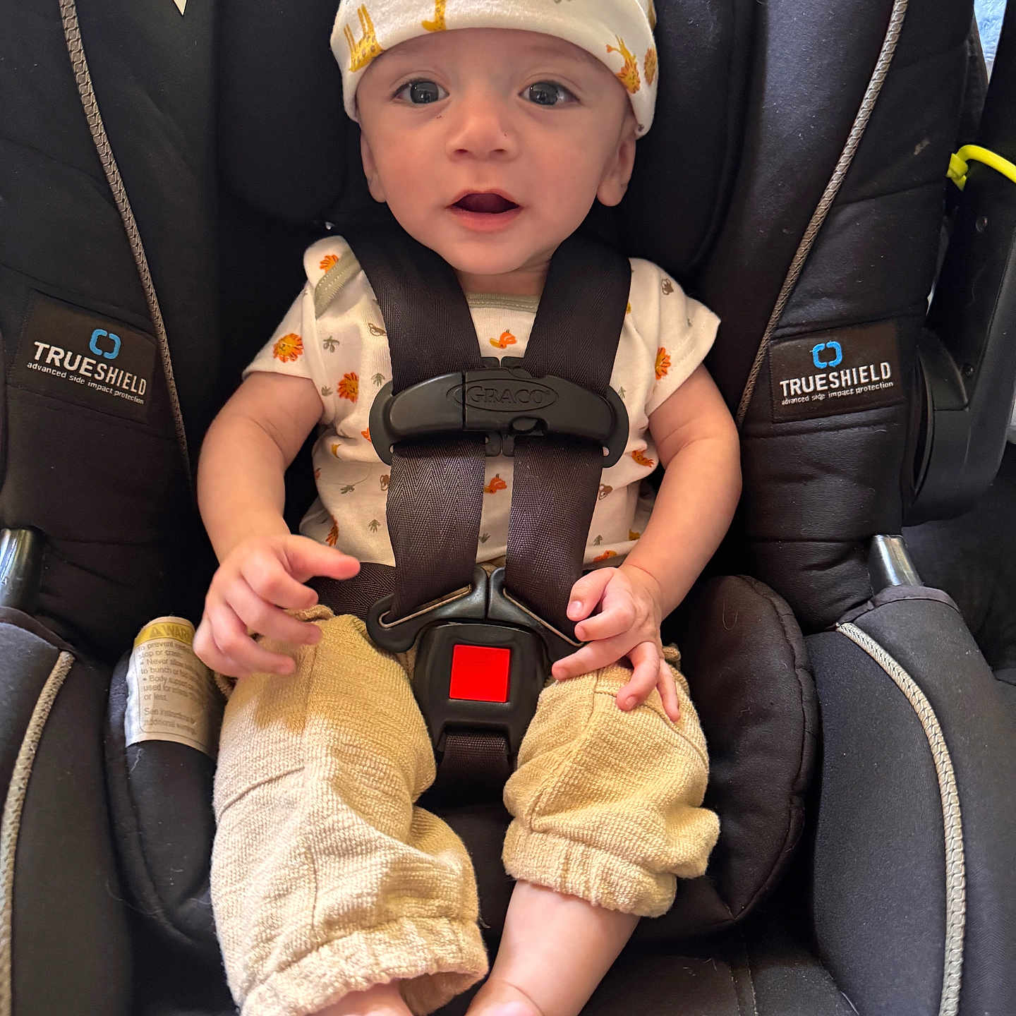 Sebastian joined the competition — help win amazing prizes! baby, child, car_seat, infant, hat, giraffe_pattern, clothing, straps, safety, seatbelt, cute, wide_eyes, open_mouth, footwear, barefoot, infant_clothing, black, yellow, beige, indoors