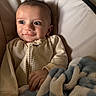 baby, infant, child, smile, face, eyes, hand, blanket, striped_clothing, onesie, cushion, soft_toy, cozy, indoors, portrait, cute, warm_lighting, relaxed, comfort, young