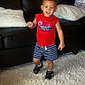 child, toddler, boy, smiling, standing, indoors, living_room, sofa, couch, pillows, rug, red_shirt, striped_shorts, sneakers, bracelet, portrait, happy, playful, race_car_shirt, home