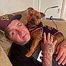 black_clothing, brown_dog, cap, casual, couch, dog, face, furniture, hand, indoor, lamp, man, pet, relaxed, ring, small_dog, star_decoration, table, tattoo, wall