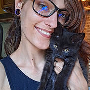 Artemis is registered to the contest to win money with this photo: person, cat, kitten, black_cat, glasses, smile, indoor, closeup, affection, pet, young_adult, earlobe_plugs, tank_top, hair, face, holding, light, window, wooden_cabinet, happy