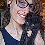 person, cat, kitten, black_cat, glasses, smile, indoor, closeup, affection, pet, young_adult, earlobe_plugs, tank_top, hair, face, holding, light, window, wooden_cabinet, happy