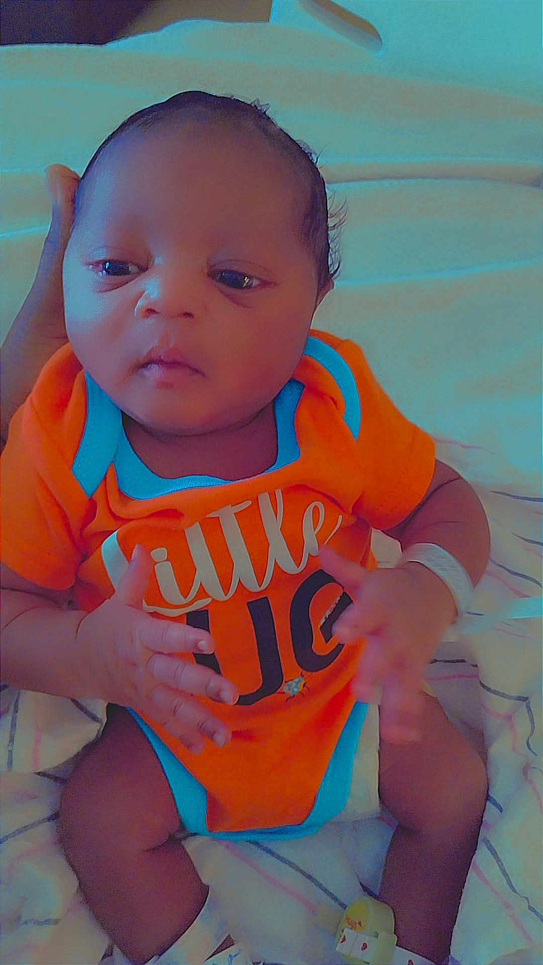 Dimitri is registered to the contest to win money with this photo: baby, infant, onesie, orange_clothing, blue_trim, blanket, hand, person, newborn, lying_down, cute, soft_lighting, indoors, sleepwear, child, face, skin, expression, small_hands, supporting_hand
