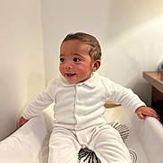 Gabriel a rejoint le concours — aidez-le/la à gagner de superbes lots ! baby, child, infant, smiling, white_clothing, onesie, sitting, indoor, soft_lighting, happy, cute, toddler, face, young_child, hands, footed_pajamas, home, cushion, portrait, playful