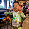child, toddler, smiling, portrait, restaurant, indoor, stuffed_toy, dinosaur_toy, gold_balloon, birthday, jacket, green_clothing, sportswear, necklace, table, booth, window, diners, background_people, happy