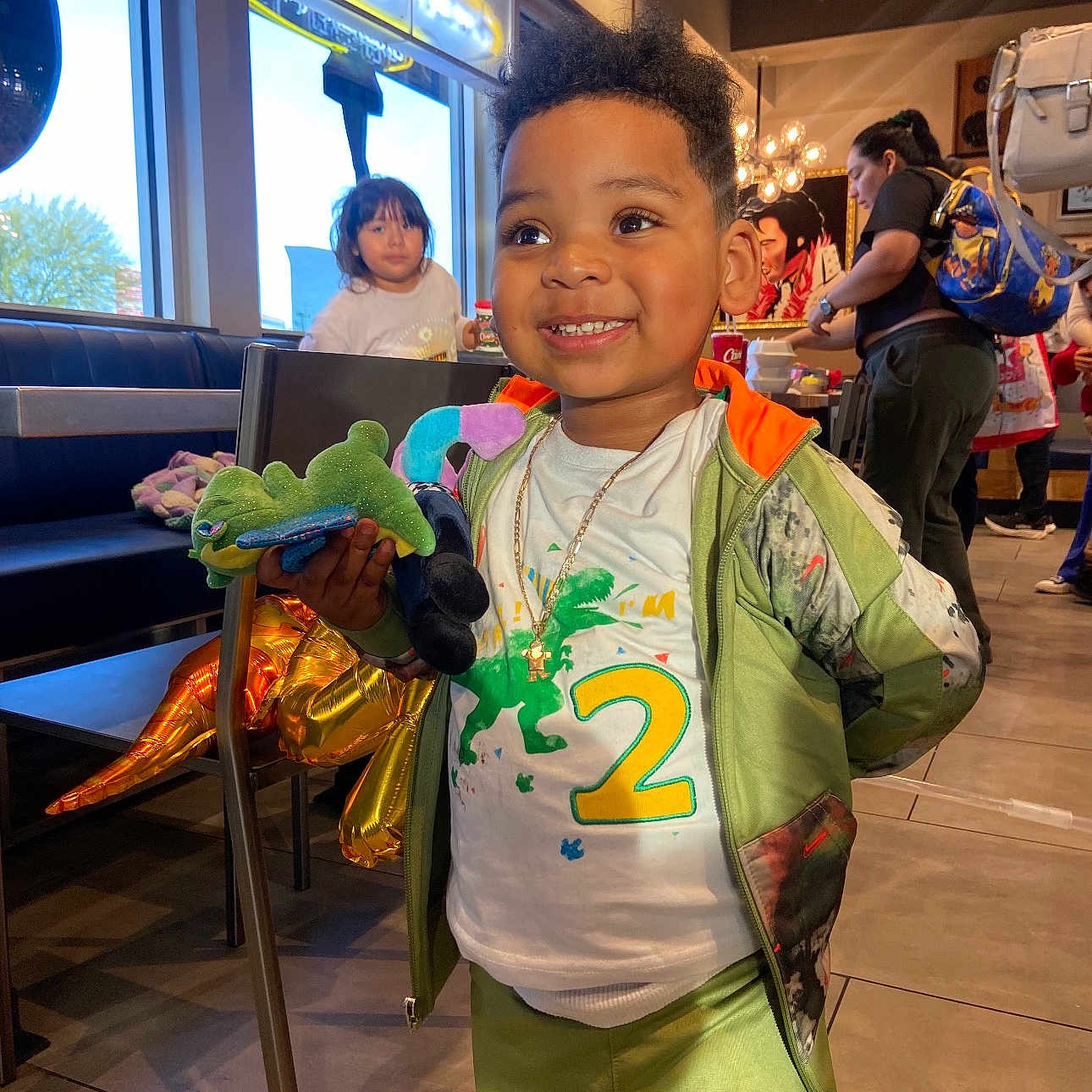 Jr joined the competition — help win amazing prizes! child, toddler, smiling, portrait, restaurant, indoor, stuffed_toy, dinosaur_toy, gold_balloon, birthday, jacket, green_clothing, sportswear, necklace, table, booth, window, diners, background_people, happy