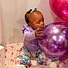 baby, child, infant, sitting, balloons, birthday, party, purple_balloon, pink_balloon, floral_clothing, lavender_shirt, hair_bows, earring, smiling, portrait, indoor, playful, celebration, blanket, colorful
