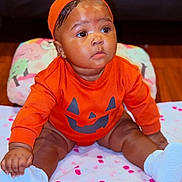 Zuri is registered to the contest to win money with this photo: baby, infant, child, portrait, sitting, orange_outfit, pumpkin_costume, halloween, hat, smiling, face, eyes, earrings, blanket, bed, indoor, shoes, white_shoes, colorful, photography