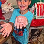 appliance_box, blue_shirt, boy, child, cooking, face, floor_tiles, food, hand, happy, home, indoor, kitchen, meatball, person, plastic_bag, plate, shorts, smiling, table