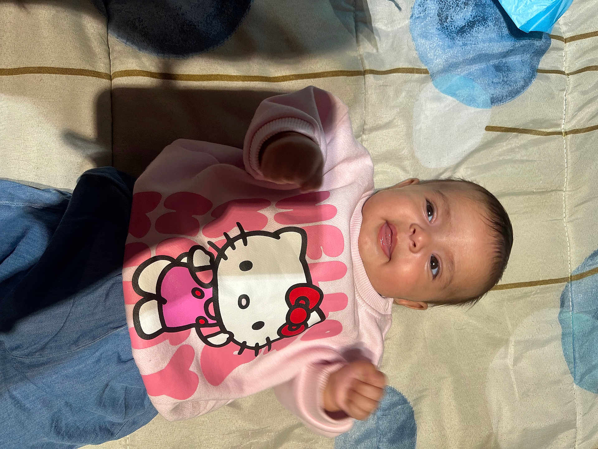 Yasmine a rejoint le concours — aidez-le/la à gagner de superbes lots ! baby, child, smiling, hello_kitty, pink_sweatshirt, blue_pants, blanket, indoor, cute, infant, portrait, happy, clothing, person, young_child, lying_down, face, head, skin, small_hands