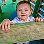 child, toddler, baby, blue_eyes, hands, wood, playground, outdoor, clothing, person, face, smiling, curious, wooden_beam, shoes, fence, colorful, play_area, young_child, daylight