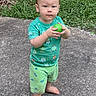 toddler, child, barefoot, green_clothing, ball, outdoor, grass, concrete, serious_expression, standing, summer, playful, cute, young_child, casual, nature, daytime, toy, shorts, tshirt
