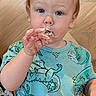 toddler, child, blue_eyes, eating, food, hand, shirt, dinosaur_print, messy, face, portrait, indoor, cute, baby, person, clothing, snack, young_child, casual, closeup