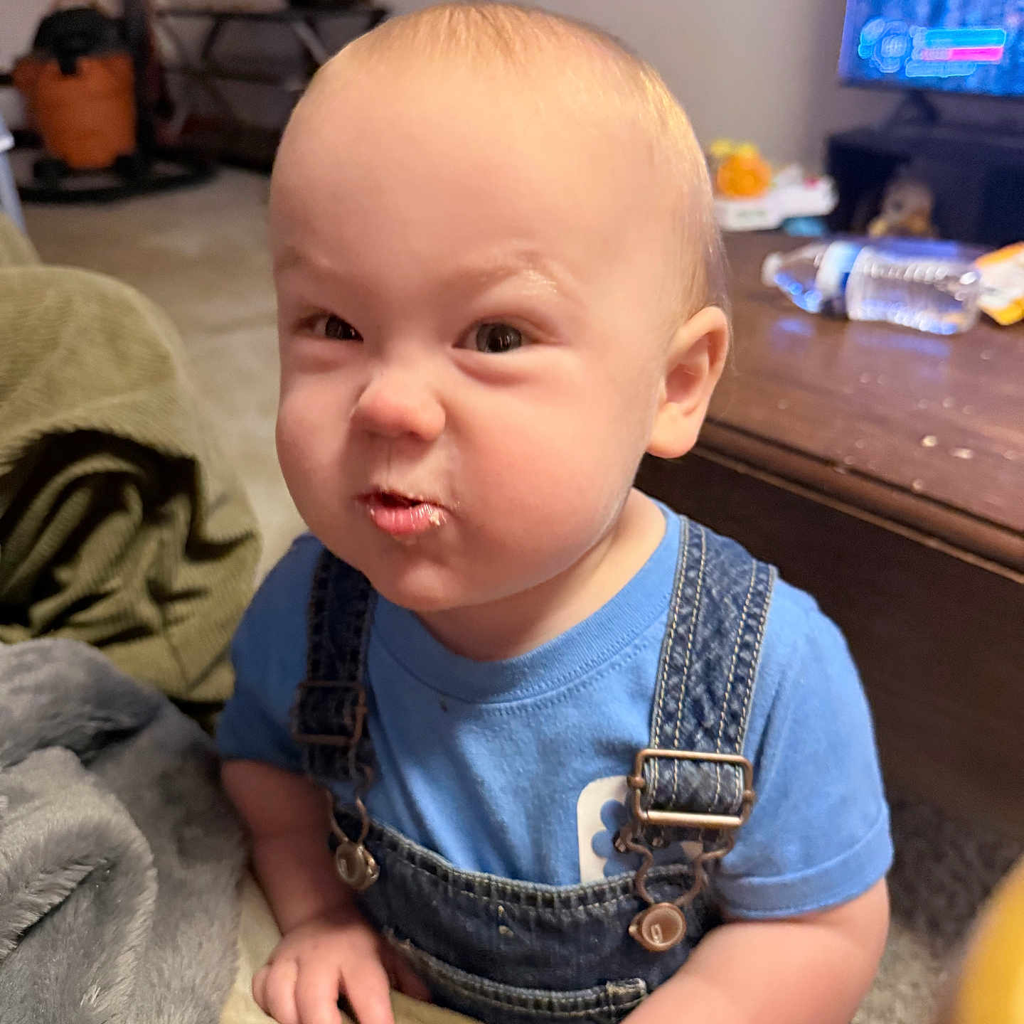 Markus joined the competition — help win amazing prizes! baby, blanket, blue_shirt, buckles, carpet, coffee_table, couch, denim_overalls, expression, funny_face, hand, indoor, infant, judgmental_look, living_room, portrait, scrunched_face, television, toddler, water_bottle