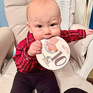 Markus is registered to the contest to win money with this photo: baby, child, infant, plaid_shirt, card, milestone, sitting, cushioned_chair, indoor, cute, portrait, chewing, hand, face, clothing, person, furniture, smile, young, celebration