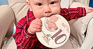 Markus is registered to the contest to win money with this photo: baby, card, celebration, chewing, child, clothing, cushioned_chair, cute, face, furniture, hand, indoor, infant, milestone, person, plaid_shirt, portrait, sitting, smile, young