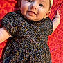 baby, blanket, child, closeup, cute, dress, fabric, face, floral_pattern, happy, headband, indoors, infant, laying_down, pattern, person, portrait, red, skin, smiling