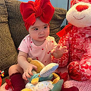 Sol is registered to the contest to win money with this photo: baby, child, red_bow, headband, onesie, toy, stuffed_animal, pink, red, couch, blanket, indoor, cute, smiling, seated, playful, soft_toys, knitted_blanket, cozy, person