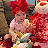 baby, child, red_bow, headband, onesie, toy, stuffed_animal, pink, red, couch, blanket, indoor, cute, smiling, seated, playful, soft_toys, knitted_blanket, cozy, person