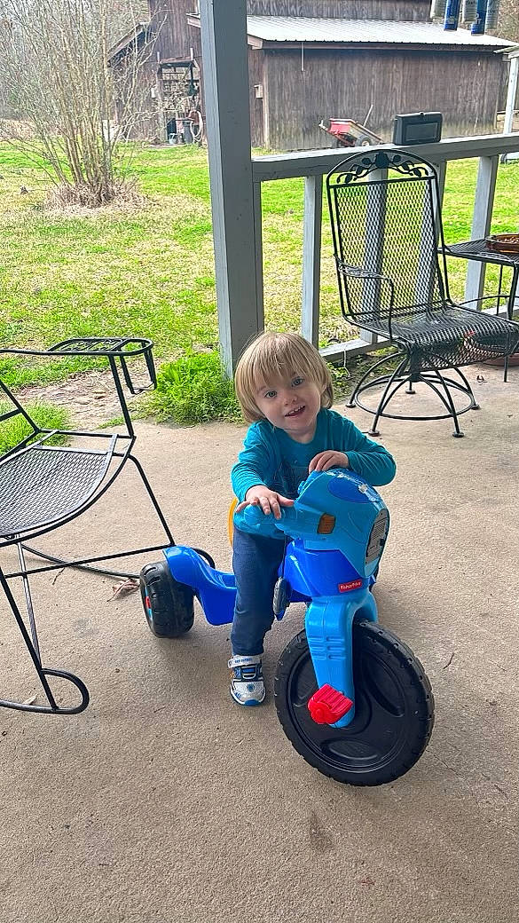 Blake is registered to the contest to win money with this photo: automotive_tire, baby, baby_toddler_clothing, chair, electric_blue, fun, grass, leisure, outdoor_furniture, person, plant, recreation, riding_toy, rolling, smile, synthetic_rubber, tire, toddler, tread, vehicle