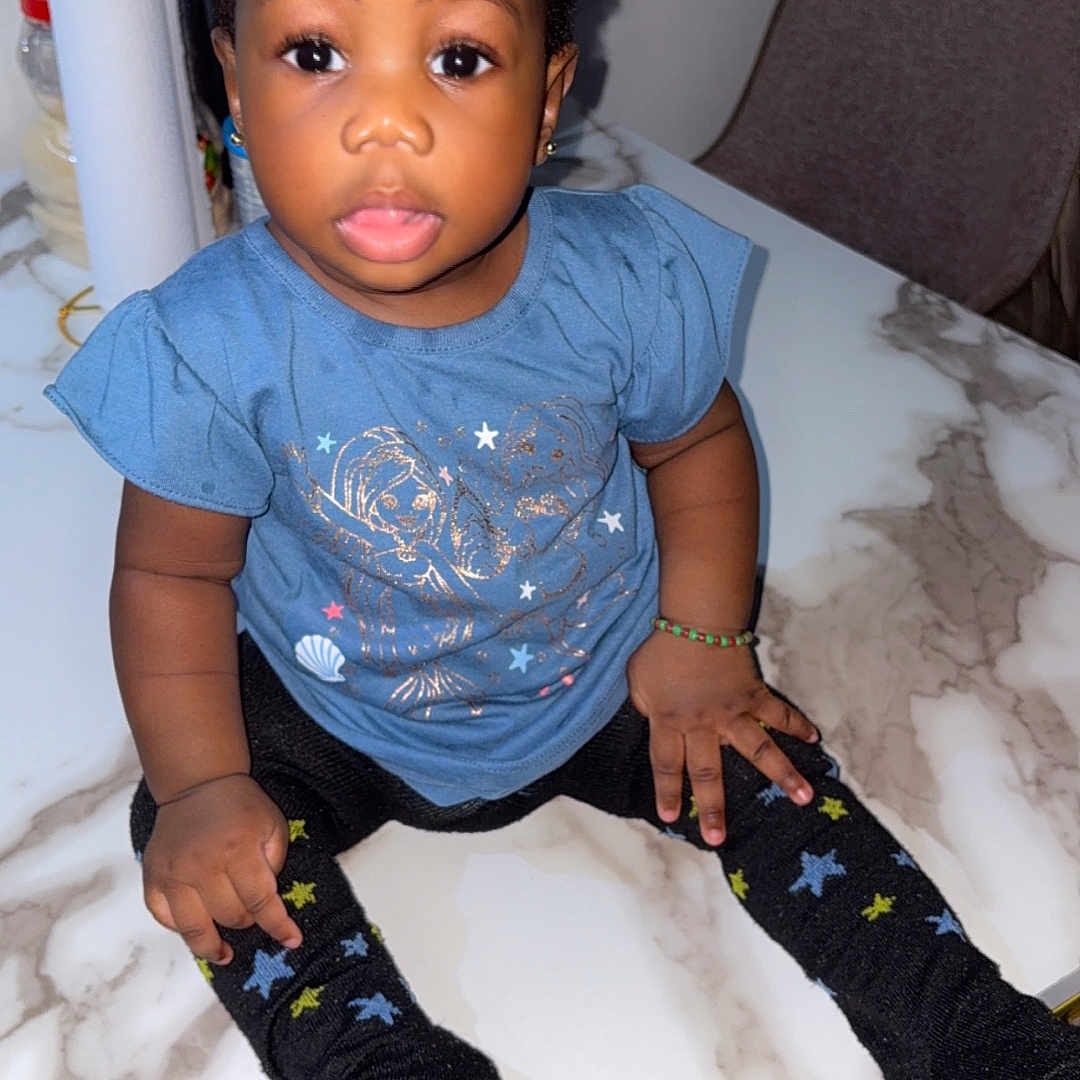 Michel is registered to the contest to win money with this photo: black_hair, black_socks, blue_shirt, bracelet, chair, child, curious_expression, curly_hair, cute, earrings, hair_buns, indoor, kitchen_items, marble_table, person, sitting, star_pattern, table, toddler, young_child