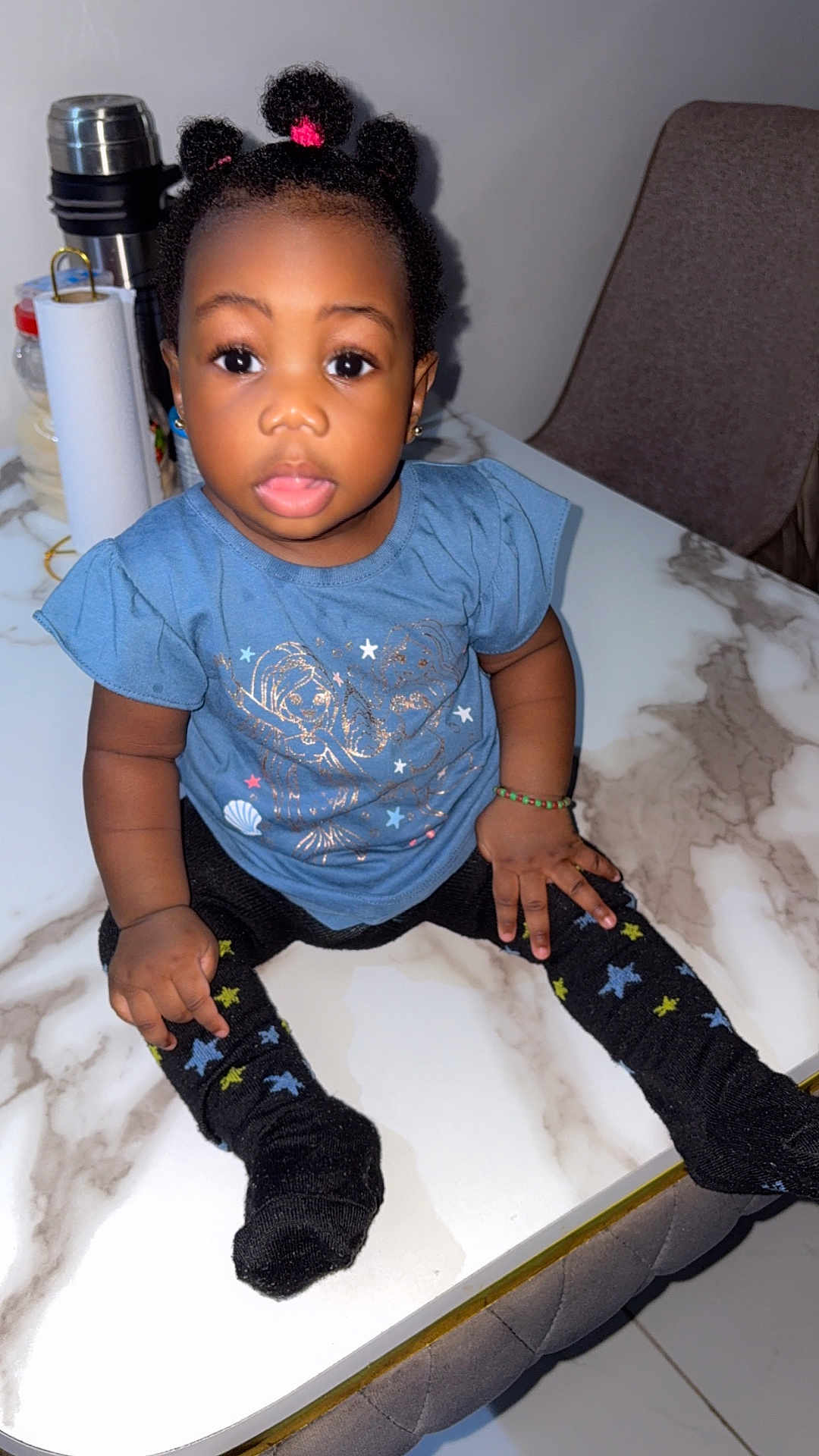Michel is registered to the contest to win money with this photo: toddler, child, blue_shirt, black_socks, star_pattern, marble_table, curly_hair, hair_buns, bracelet, earrings, indoor, chair, kitchen_items, curious_expression, sitting, person, young_child, cute, table, black_hair