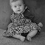 Violet is registered to the contest to win money with this photo: baby, child, infant, leopard_print, dress, sitting, carpet, cute, portrait, indoor, face, smiling, person, clothing, long_sleeve, barefoot, young, expression, curly_hair, black_and_white