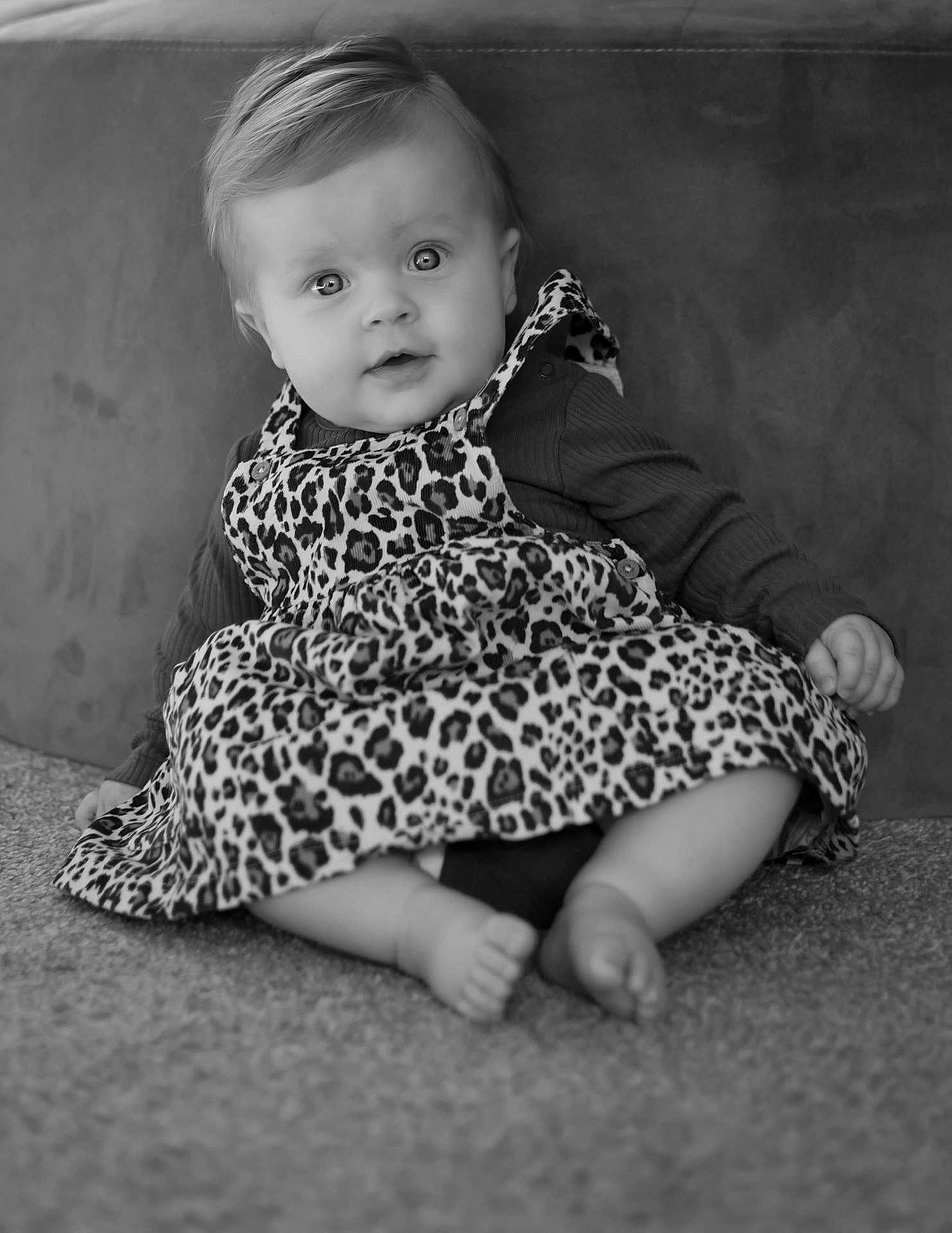 Violet is registered to the contest to win money with this photo: baby, child, infant, leopard_print, dress, sitting, carpet, cute, portrait, indoor, face, smiling, person, clothing, long_sleeve, barefoot, young, expression, curly_hair, black_and_white