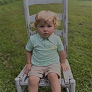 Logan joined the competition — help win amazing prizes! toddler, child, boy, curly_hair, blonde, green_grass, outdoor, chair, wooden_chair, rustic, polo_shirt, shorts, serious_expression, nature, field, summer, sitting, person, portrait, daylight