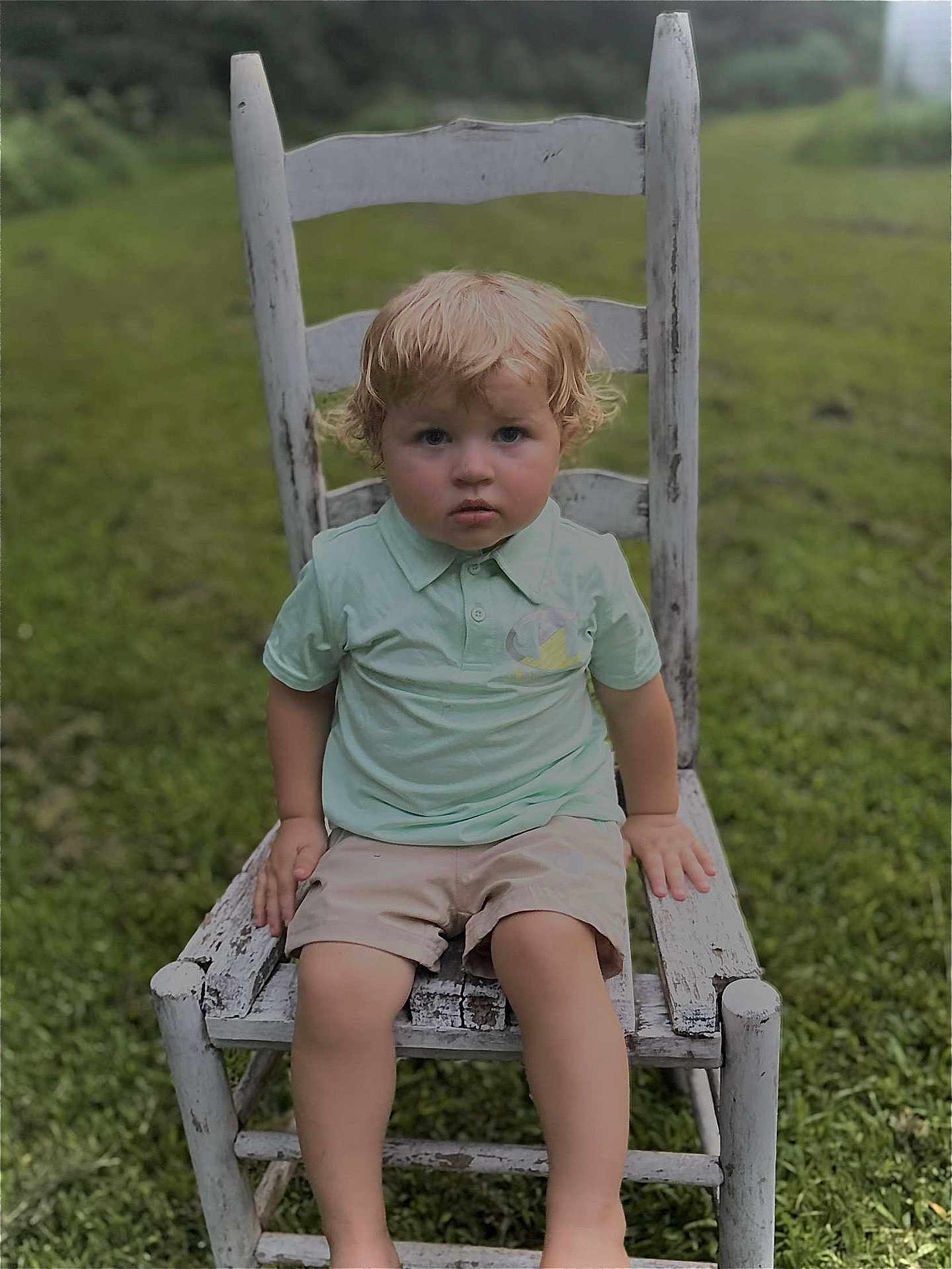 Logan joined the competition — help win amazing prizes! toddler, child, boy, curly_hair, blonde, green_grass, outdoor, chair, wooden_chair, rustic, polo_shirt, shorts, serious_expression, nature, field, summer, sitting, person, portrait, daylight