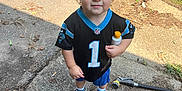 Logan is registered to the contest to win money with this photo: toddler, child, person, outdoor, daylight, jersey, sportswear, football, bottle, shoes, hose, driveway, vehicle, car, red_sled, greenery, sidewalk, curly_hair, expression, concrete