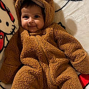 Nevaeh is registered to the contest to win money with this photo: baby, child, smiling, onesie, brown_clothing, fluffy, cute, portrait, indoor, blanket, cartoon, soft_texture, face, headwear, cozy, infant, warm_clothing, happy, lying_down, adorable