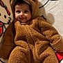 baby, child, smiling, onesie, brown_clothing, fluffy, cute, portrait, indoor, blanket, cartoon, soft_texture, face, headwear, cozy, infant, warm_clothing, happy, lying_down, adorable