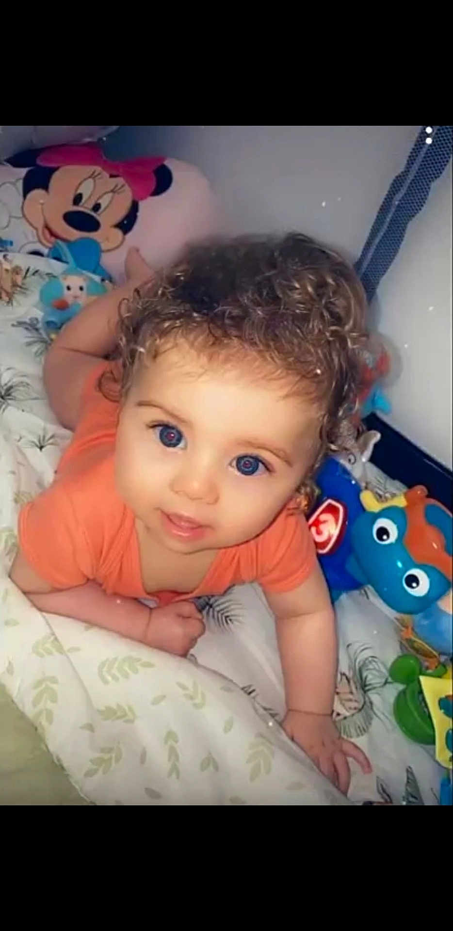 Menzo participe au concours pour gagner de l'argent avec cette photo : baby, child, curly_hair, blue_eyes, toys, minnie_mouse, pillow, bed, blanket, orange_shirt, infant, cute, indoor, playful, person, smiling, lying_down, colorful, soft, closeup