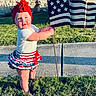 blue, child, cute, flag, grass, headwrap, holding, outdoor, patriotic, person, plaid, portrait, red, ribbon, sidewalk, skirt, smile, sunlight, toddler, white