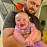 baby, man, beard, purple_tutu, smile, laughing, indoor, staircase, living_room, holding, affection, happy, person, child, family, casual_clothing, home, cute, human, portrait