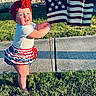 baby, blue, brick_wall, building, car, child, curious, flag, grass, happy, headwrap, outdoor, patriotic, red, sidewalk, skirt, standing, sunlight, white, window