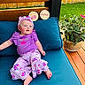 baby, barefoot, bench, child, cushion, floral_pattern, flower_pot, grass, happy, headband, infant, milestone_sign, natural_light, outdoor, plant, portrait, purple_clothing, sitting, smiling, wooden_fence