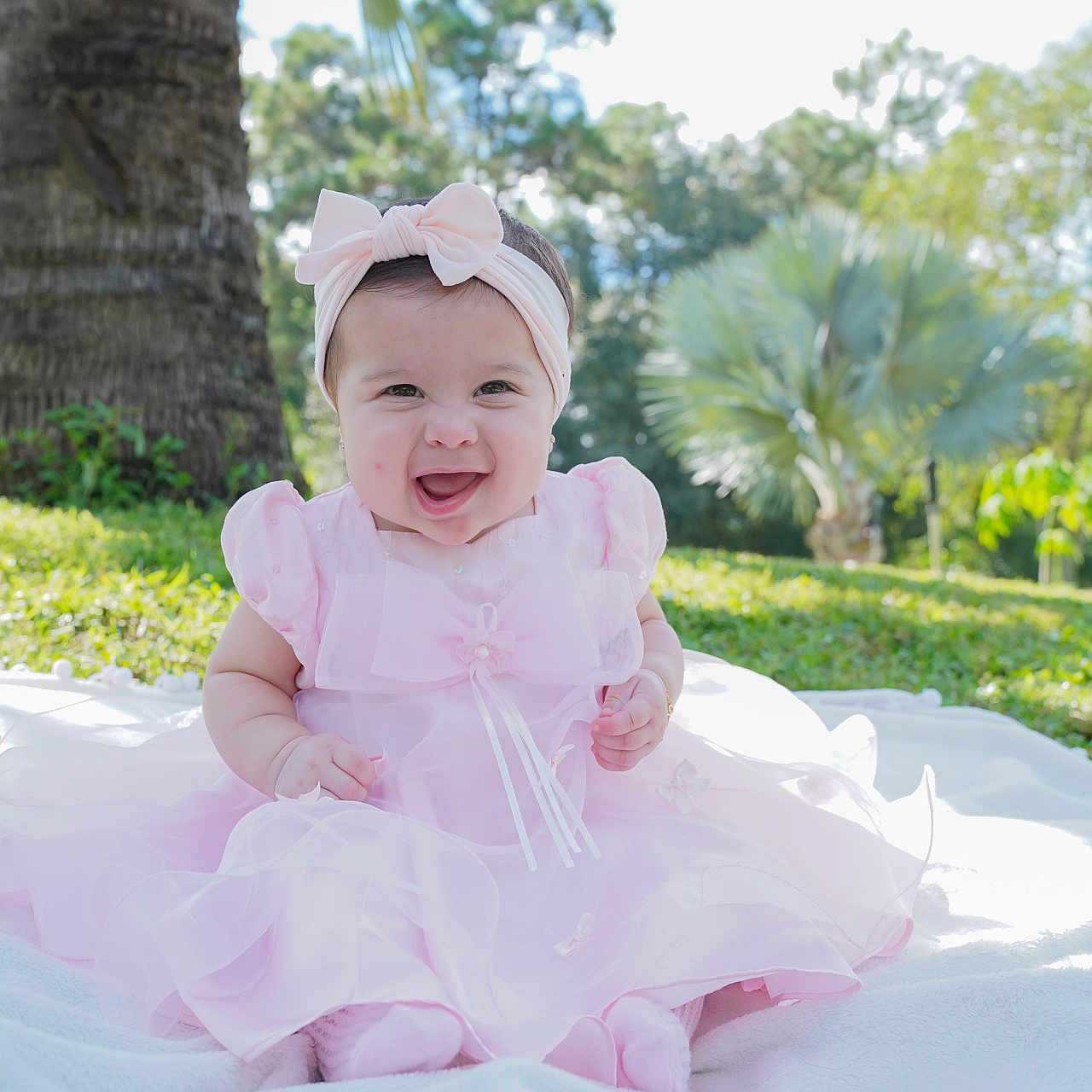 Melody is registered to the contest to win money with this photo: baby, child, infant, smiling, pink_dress, headband, bow, outdoor, garden, palm_tree, tropical, portrait, sitting, blanket, grass, sunlight, nature, happy, dress, family_photo