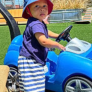 Aureliano joined the competition — help win amazing prizes! toddler, child, toy_car, blue_hat, striped_shorts, barefoot, outdoor, grass, playground, steering_wheel, sunny, summer, casual_clothing, plastic_toy, young_child, daytime, greenery, fun, curious, park