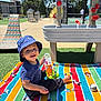Aureliano is registered to the contest to win money with this photo: toddler, child, smiling, hat, blue_clothing, toy, blanket, outdoor, sunny, playtime, water_table, colorful, striped_blanket, mickey_mouse, green_grass, plastic_toys, play_area, happy, sitting, daytime