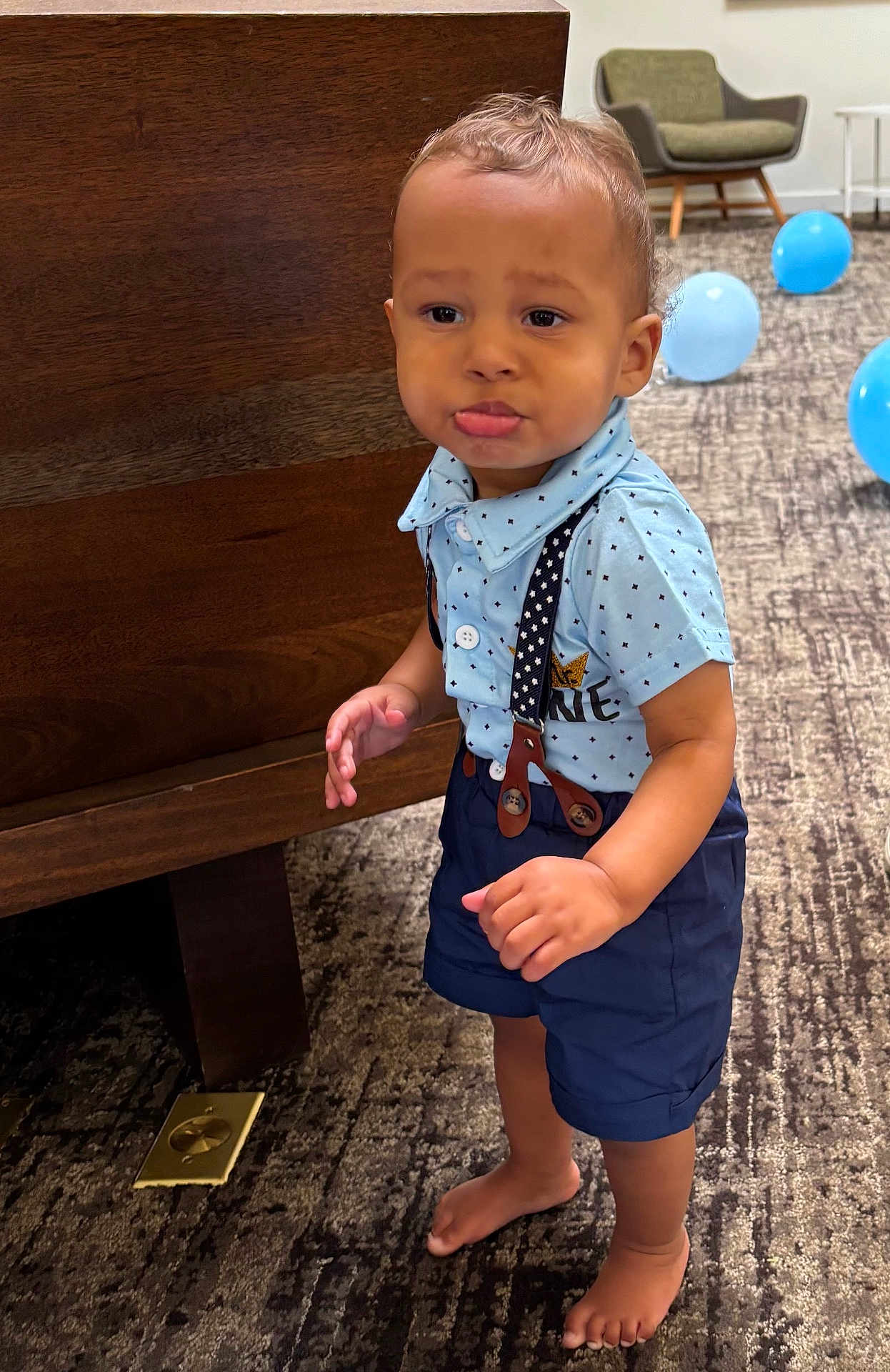 Aureliano is registered to the contest to win money with this photo: toddler, child, barefoot, blue_shirt, suspenders, navy_shorts, carpet, wooden_furniture, balloons, indoor, pout, standing, cute, expression, modern_chair, playful, person, clothing, floor, room
