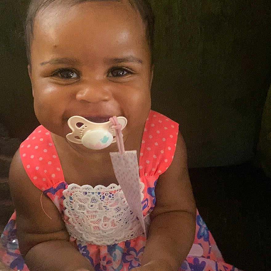 Dalaney is registered to the contest to win money with this photo: accessory, baby, child, colorful, couch, cute, dress, face, happy, holding, indoor, pacifier, person, pink_bow, plug_adapter, portrait, sitting, smile, toddler, young_child