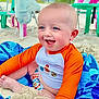 baby, child, beach, sand, blue_eyes, orange_shirt, towel, smiling, happy, sitting, outdoor, summer, vacation, playful, sunny, sand_play, footwear_free, daytime, person, casual