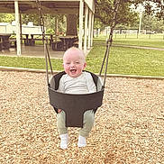 Heston is registered to the contest to win money with this photo: baby, swing, playground, child, grass, woodchips, trees, picnic_table, smiling, happy, outdoor, park, daylight, clothing, sock, seat, metal_chain, nature, person, fun