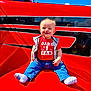 baby, child, red_shirt, jeans, socks, smiling, outdoor, sunny, blue_sky, clouds, red_surface, casual_clothing, happy, portrait, sitting, cute, young_child, daylight, face, person