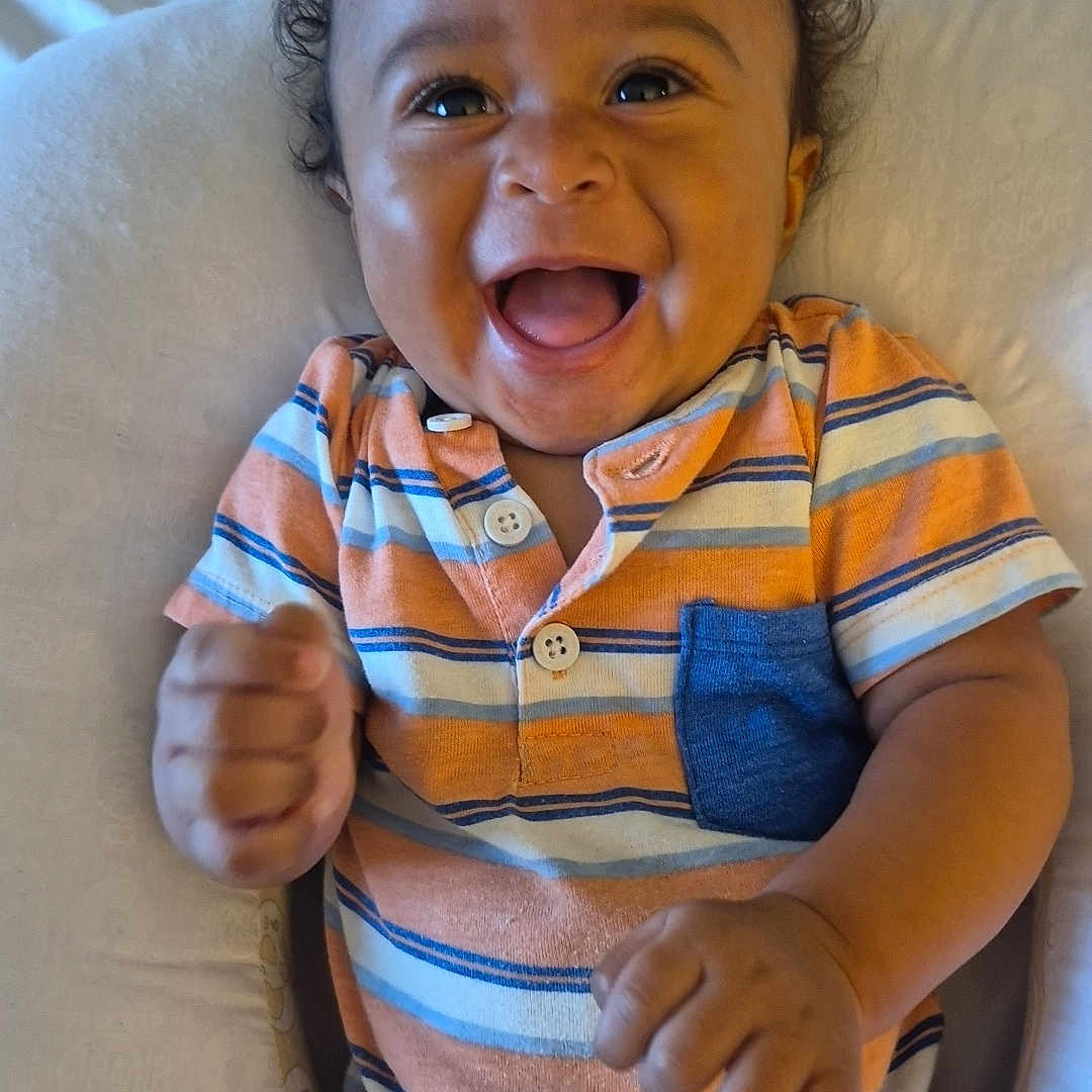 Brooks joined the competition — help win amazing prizes! baby, child, infant, smiling, laughing, curly_hair, portrait, sitting, striped_clothing, orange_clothing, blue_details, onesie, happy, joyful, indoor, cushion, seat, close_up, hands, family_photo